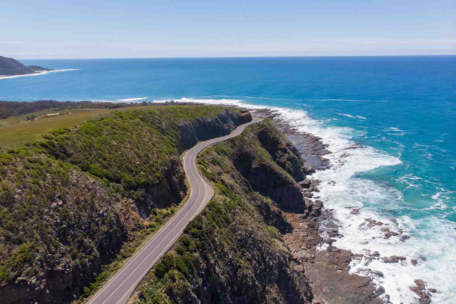 Imagem de uma estrada com um morro ao lado esquerdo e do direito o mar, vistos de cima de dia 
