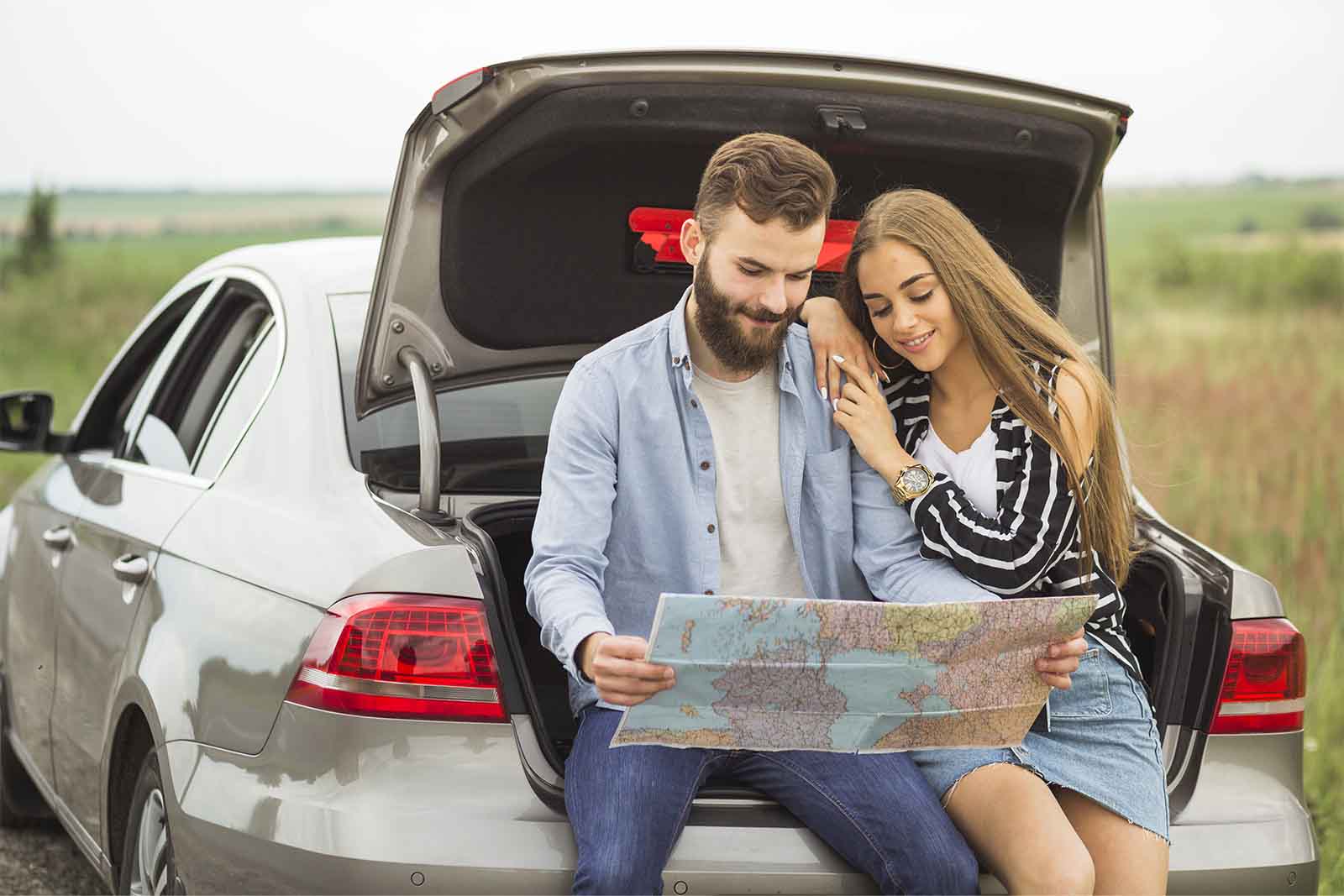 Imagem de um casal sentado no porta malas aberto de um carro alugado, ambos olhando um mapa impresso, na beira de estrada, de dia 