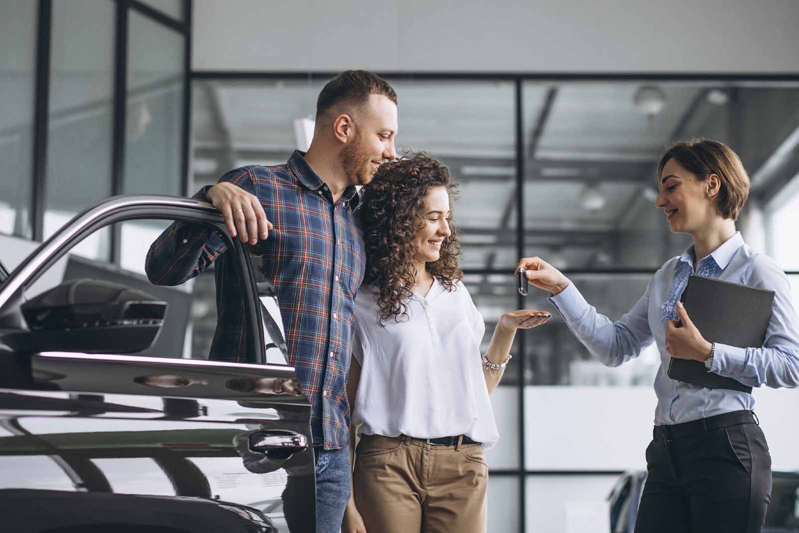 Imagem de um casal recebendo a chave do carro de uma vendedora, sobre um carro que acabaram de alugar, dentro de uma concessionaria de dia 