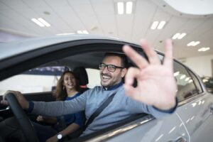 Imagem de um casal dentro de um carro em um concessionaria, acabando de alugar um carro com sua esposa sentada no banco de passageiro