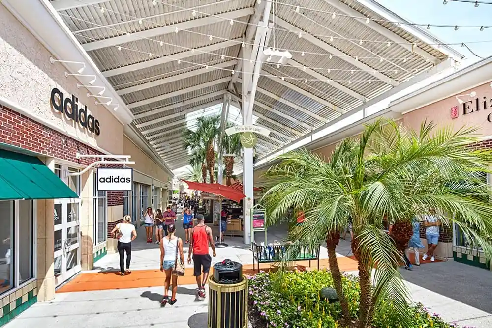 Shopping outlet ao ar livre com palmeiras e lojas, como Adidas, onde pessoas fazem compras. Destino de viagem ideal para lazer e experiências de compras em clima tropical.