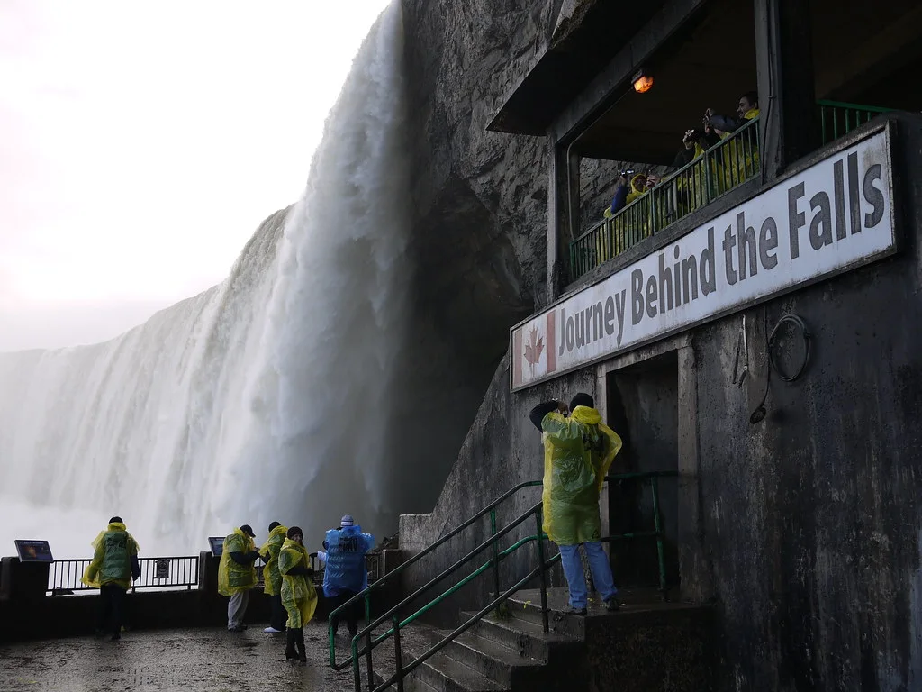 Turistas com capas de chuva amarelas na atração "Journey Behind the Falls" das Cataratas do Niágara, Canadá. A imagem mostra a grandiosa queda d'água e a estrutura de visitação com plataforma de observação.