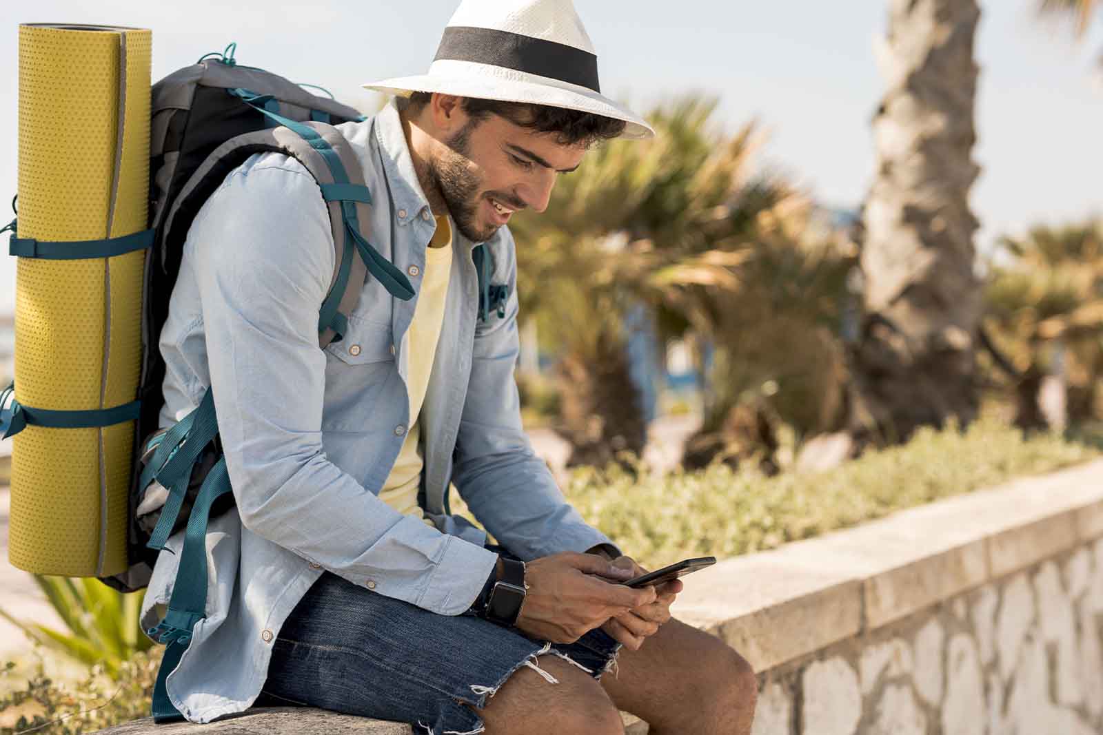 Imagem de um turista sentado em um parapeito com chapéu, segurando um celular olhando para o mesmo de dia 