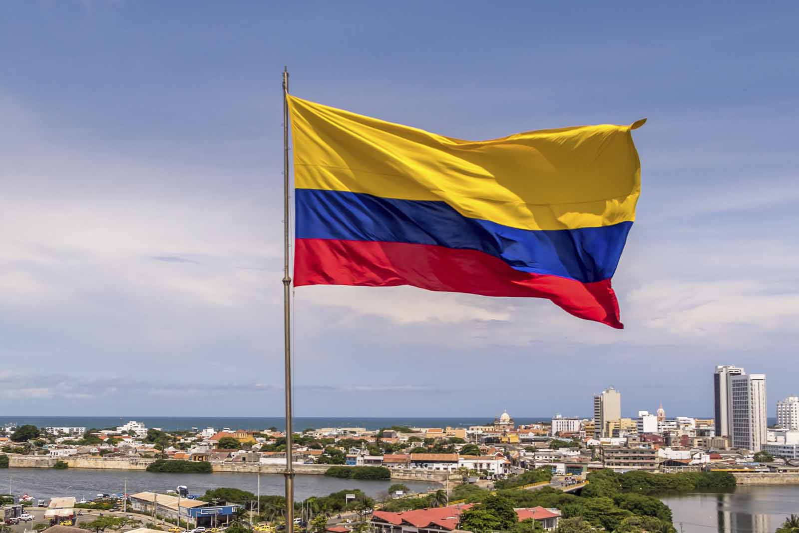 Imagem da cidade de Cartagena na Colômbia, com a bandeira da Colômbia no centro da imagem e ao fundo a cidade com diversos edifícios, árvores, lagos e de dia 