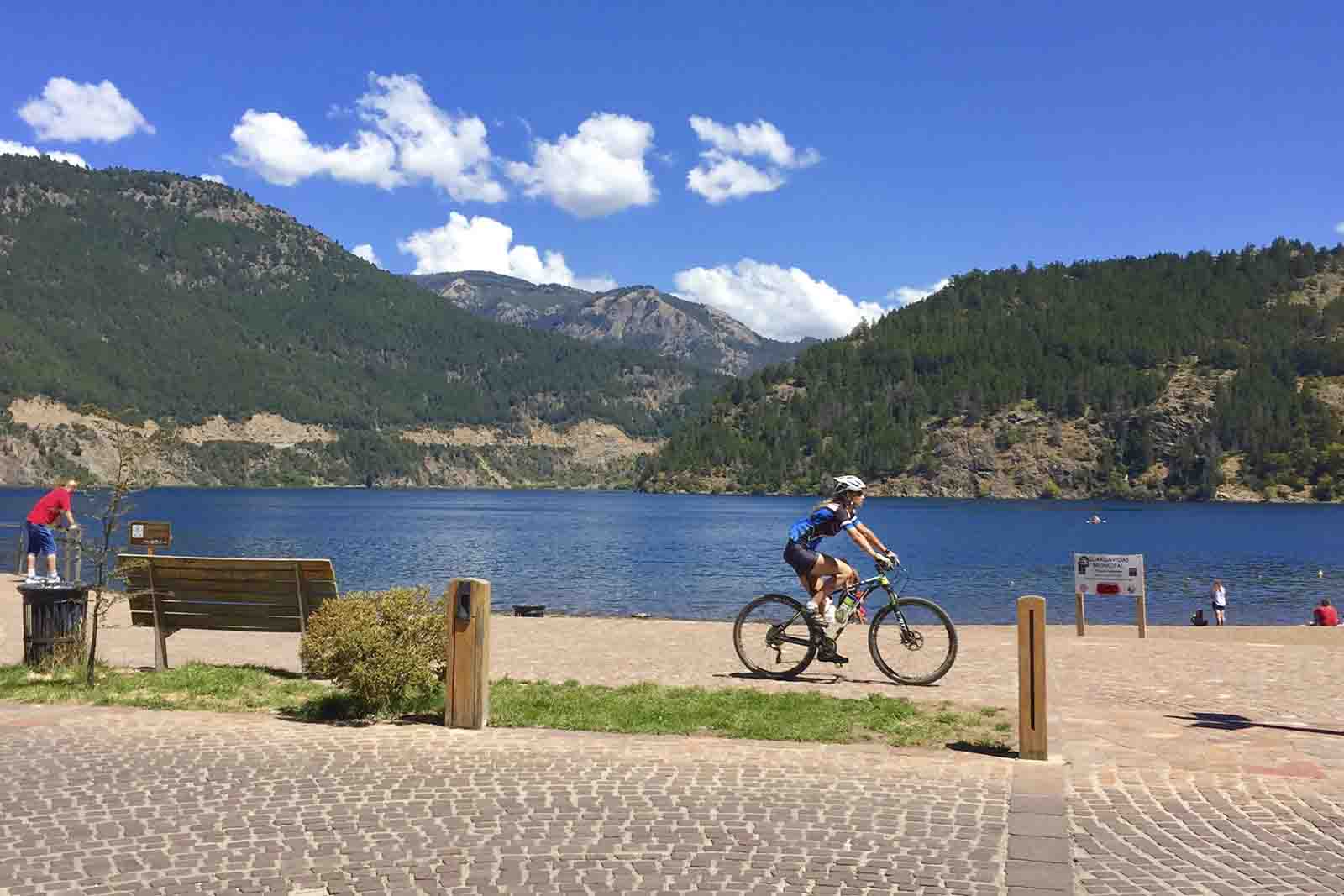 Imagem de Patagônia na Argentina, com uma praça na beira do lago, pessoas transitando a pé e de bicicleta, com o fundo de montanhas e nuvens de dia