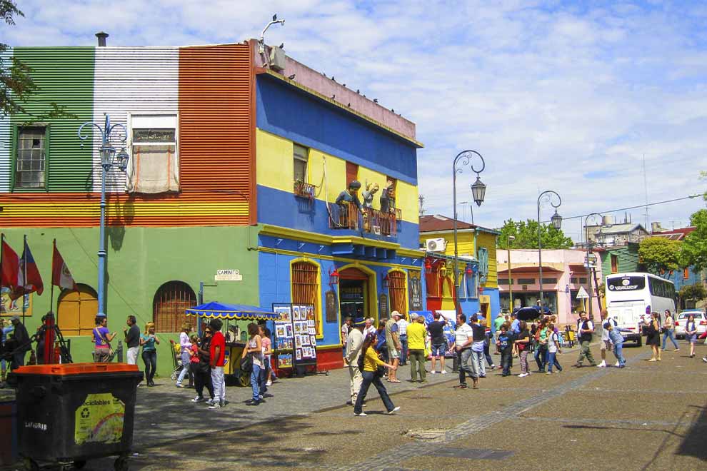 Bairro La Boca, com diversas construções coloridas e pessoas, durante o dia, com o céu azul