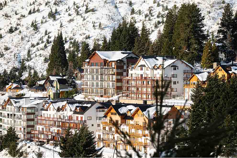 Bariloche no inverno, com prédios, árvores e montanhas cobertas de neve, durante o dia. 