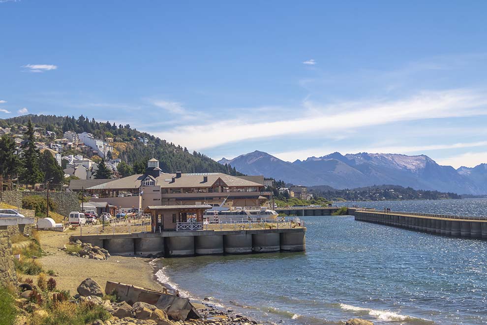 Cidade de Bariloche, no Porto de São Carlos, com casas na beira do mar, com prédios e montanhas ao fundo, durante o dia. 