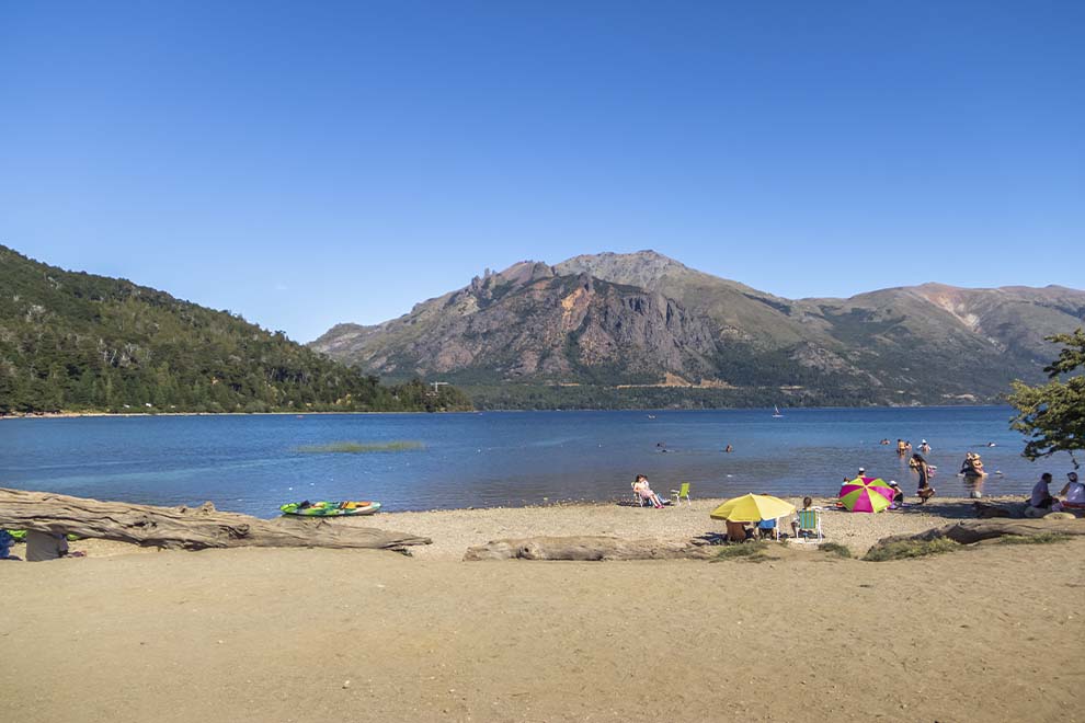 Bariloche, no lago gutierrez, areia na beira do lago com pessoas, montanhas ao fundo, durante o dia. 