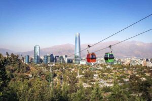 Cidade de Santiago no Chile, com diversos prédios e árvores ao fundo, com duas linhas de teleférico e de dia