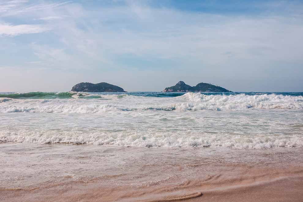 Praia com ondas e ilhas ao fundo em dia ensolarado